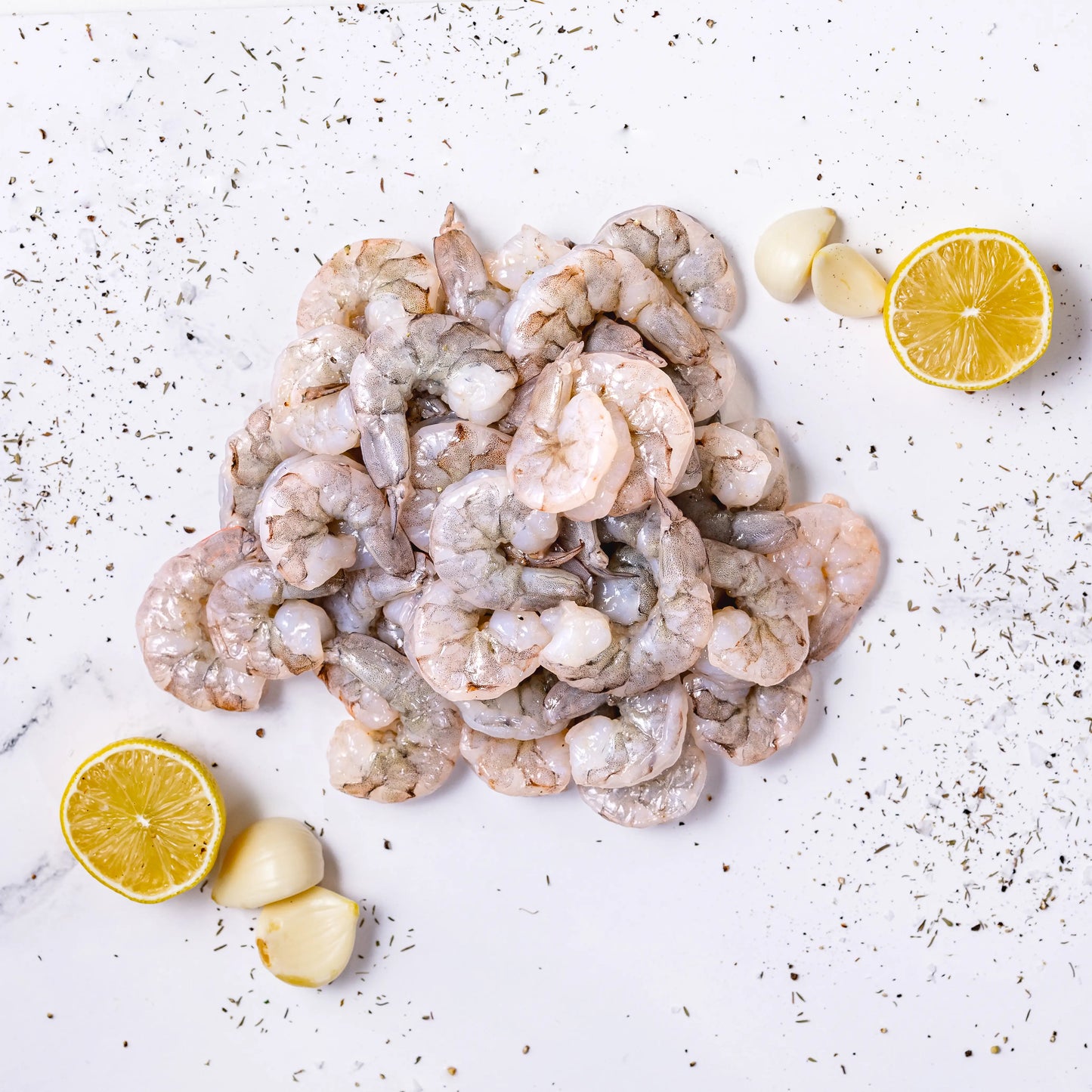 Wild-Caught Shrimp arranged on a white surface, accented with garlic cloves, two lemon halves, and a sprinkle of dried herbs.