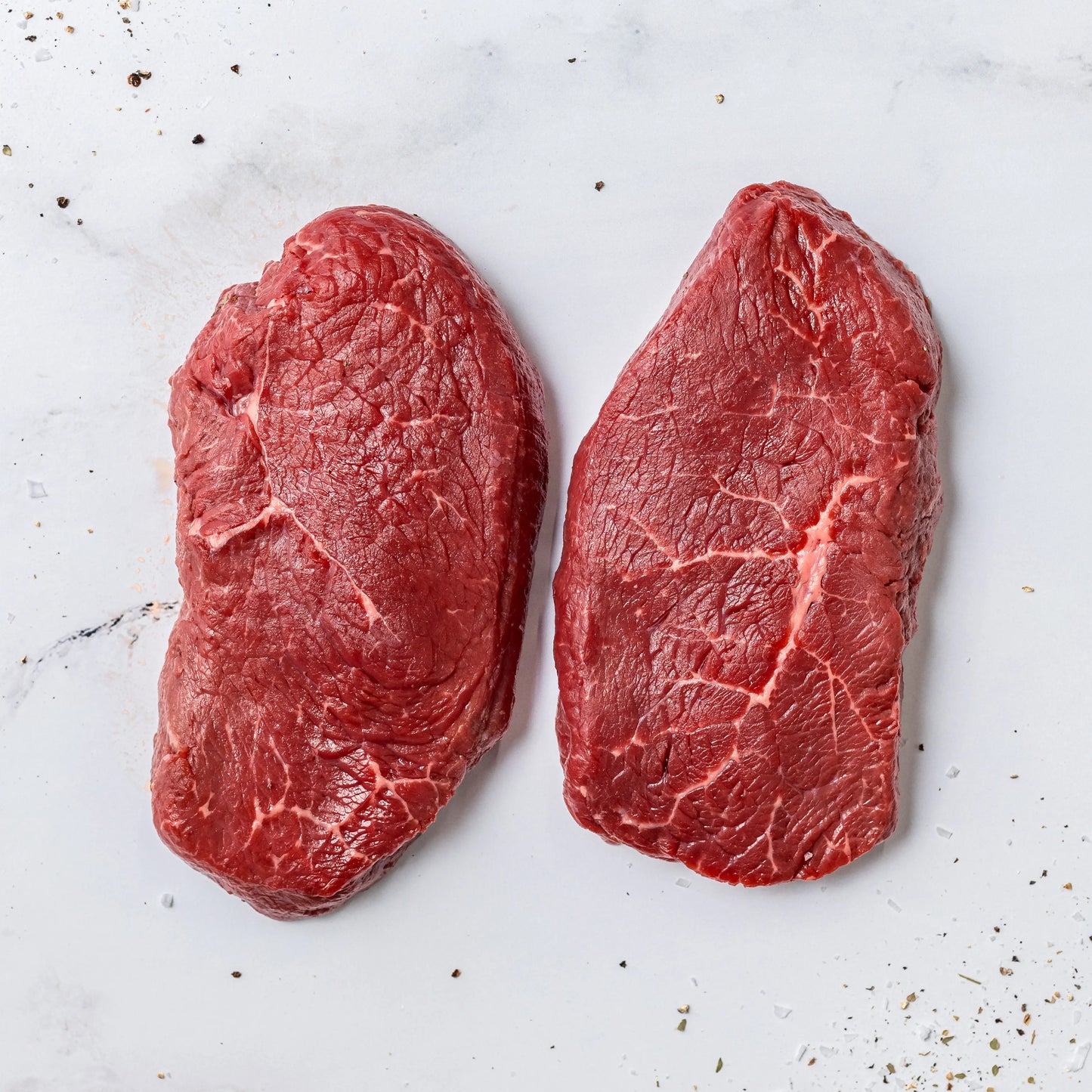 Two Steak Cut Sirloin Steaks on a white marble surface, with scattered grains of salt and black pepper around them.