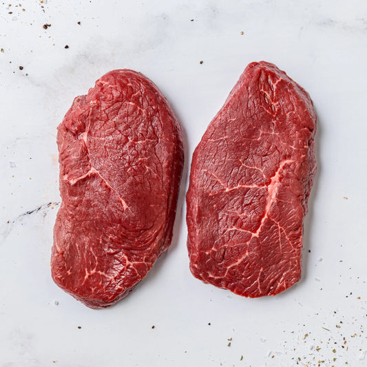 Two Steak Cut Sirloin Steaks on a white marble surface, with scattered grains of salt and black pepper around them.