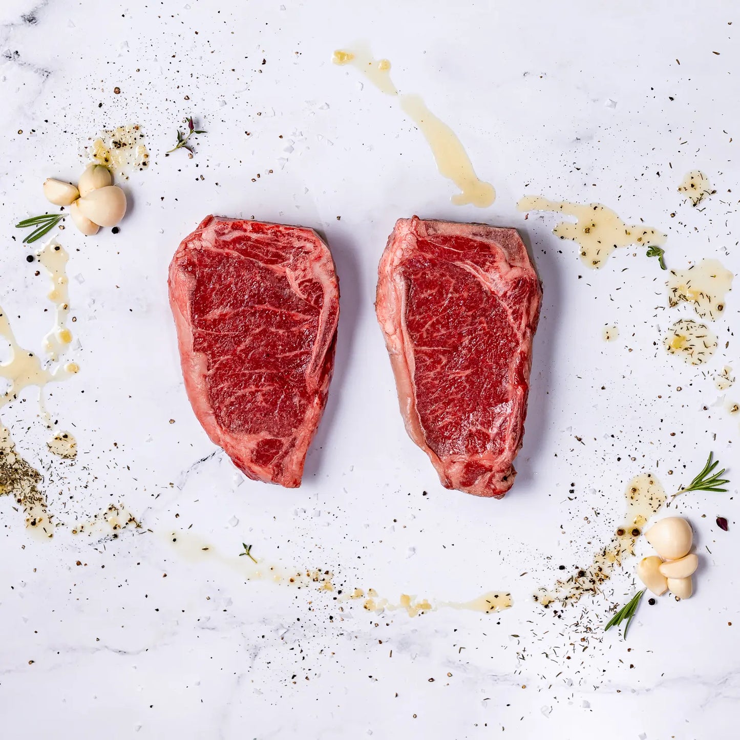 Two raw bone-in New York strip steaks on a white surface with seasonings, herbs, and garlic around them.