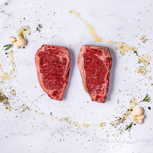 Two raw bone-in New York strip steaks on a white surface with seasonings, herbs, and garlic around them.